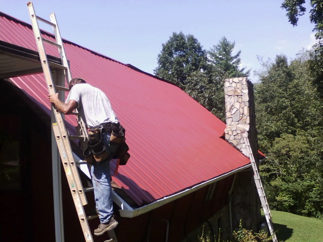 Roofer on a ladder beside a red metal roof and stone chimney — Soffit & Fascia Repair by Roofing Brothers in Ellabell